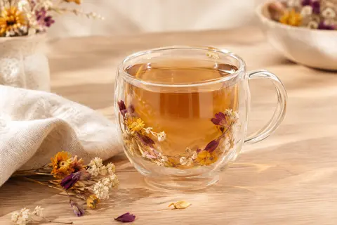 Tasse transparente à double paroi avec fleurs séchées, posée sur une table en bois clair.