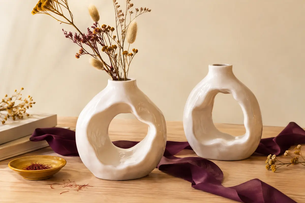 Deux vases décoratifs ronds en céramique blanche ajourée, présentés sur une table en bois clair.