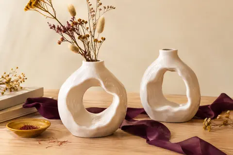 Deux vases décoratifs ronds en céramique blanche ajourée, présentés sur une table en bois clair.