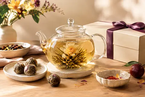 Théière en verre avec une fleur de thé blanc épanouie, accompagnée de boules de thé et d’un coffret cadeau.