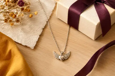Collier en acier inoxydable avec médaillon cœur à ailes d’ange et chaîne fine posé sur un fond crème.