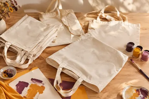 Cinq tote bags en coton naturel, unis et pliables, présentés avec du matériel de décoration textile.