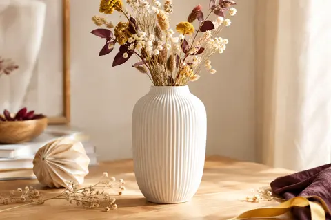 Vase décoratif blanc mat en céramique à reliefs verticaux, posé sur une table en bois clair avec des tiges séchées.