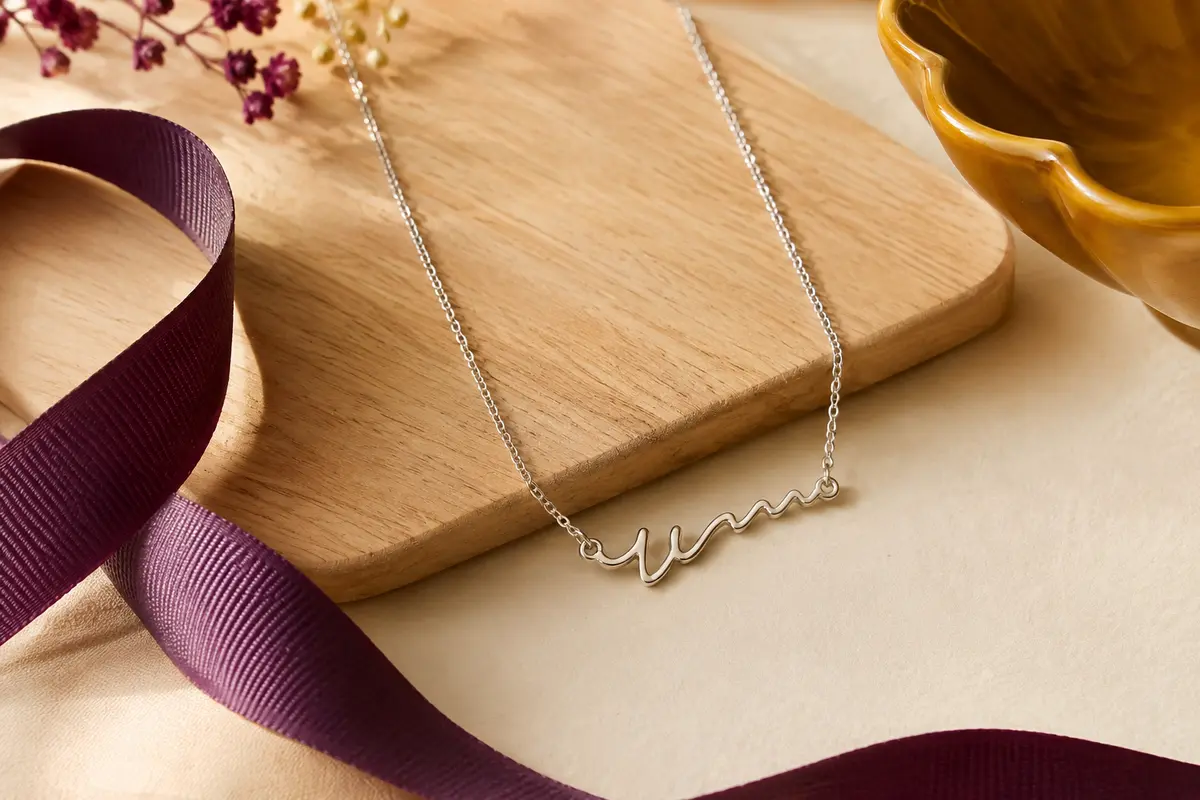 Collier prénom en argent sterling 925 à pendentif cursif, présenté sur un fond crème avec un ruban prune.