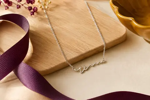 Collier prénom en argent sterling 925 à pendentif cursif, présenté sur un fond crème avec un ruban prune.