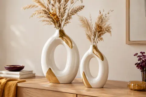 Deux vases ovales creux en céramique blanche et dorée posés sur une console en bois clair.