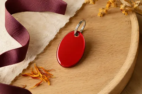 Médaille ovale rouge en aluminium avec anneau, posée sur un fond crème et bois clair.