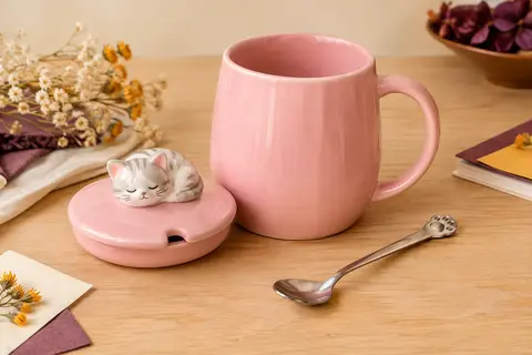 Mug rose en céramique à couvercle chat, avec une cuillère posée sur un fond crème et bois clair.