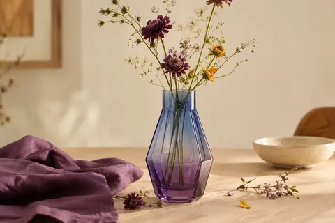 Vase géométrique en verre dégradé bleu violet posé sur une table en bois clair avec quelques fleurs.