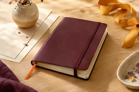 Carnet souple lie de vin avec élastique, ruban orange et pages ivoire sur un fond de bois clair.