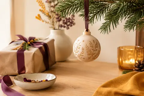 Boule de Noël ronde en tilleul clair avec ruban, présentée près de branches de sapin sur un fond crème.