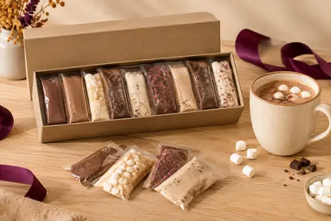 Coffret de neuf mélanges pour chocolat chaud présenté près d’une tasse en céramique et de guimauves