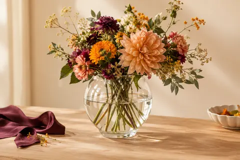 Vase Zinnia en verre transparent, forme calice, garni de tiges florales sur une table en bois clair.