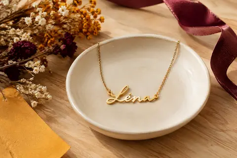 Collier prénom personnalisé à chaîne fine et pendentif doré posé sur un décor crème et bois clair.