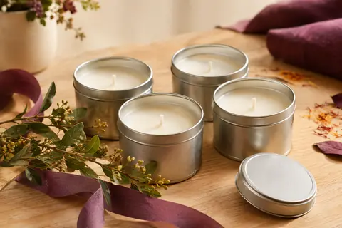Quatre bougies parfumées en pots métalliques, présentées sur un fond crème et bois clair.