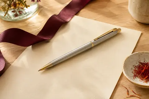 Stylo bille en acier brossé aux attributs dorés, posé sur du papier crème et du bois clair