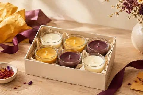 Six bougies votives parfumées en pots de verre, disposées sur un fond crème et bois clair.