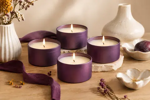 Quatre bougies parfumées en contenants métalliques violets présentées sur un fond crème et bois clair.